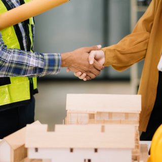 Two people shaking hands over a table of blueprints and a yellow hard hat. One person is in a hi-viz vest and the other is in office-wear