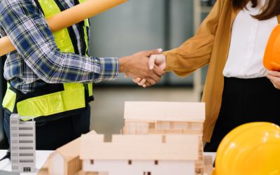 Two people shaking hands over a table of blueprints and a yellow hard hat. One person is in a hi-viz vest and the other is in office-wear
