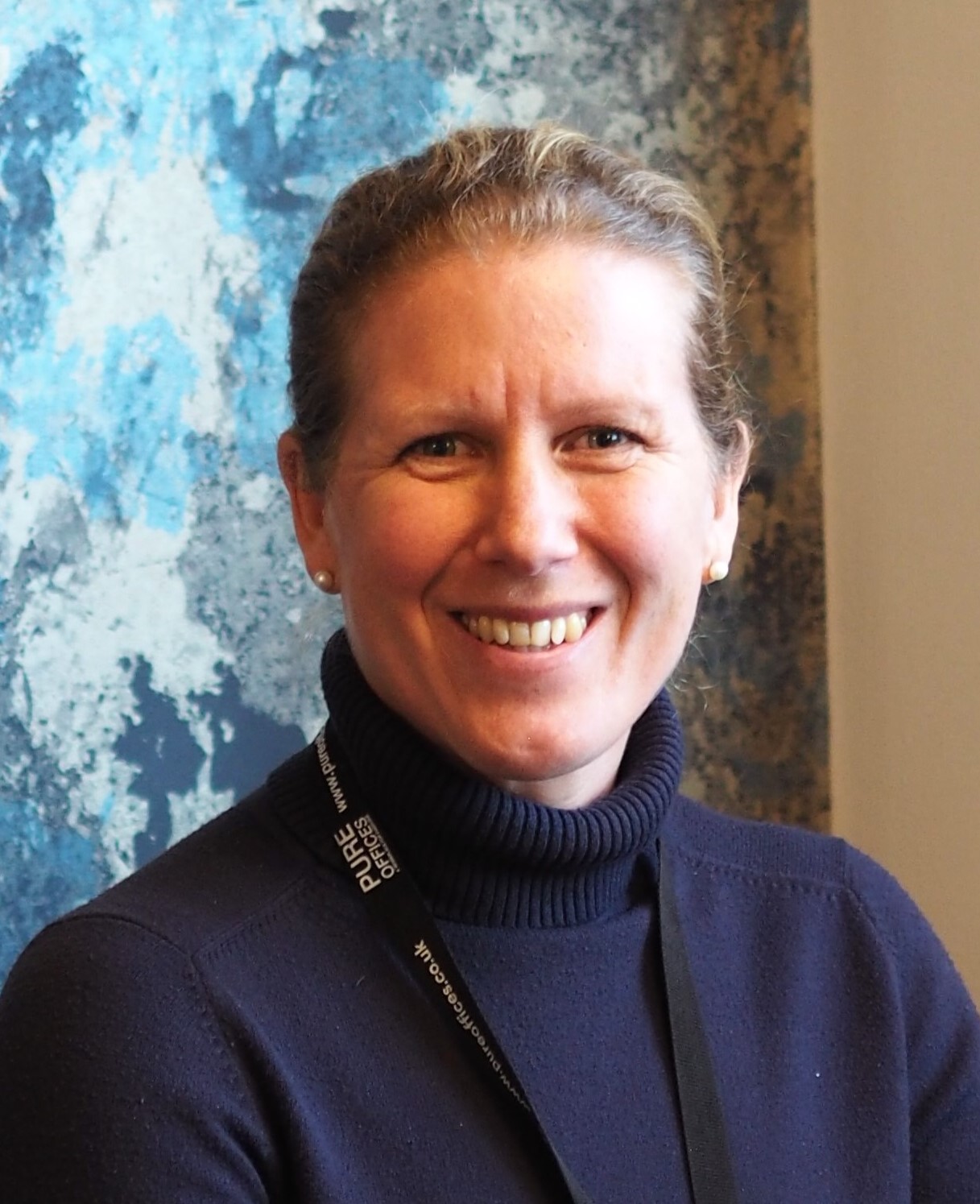 A woman with blonde hair tied back and wearing a navy blue turtle-neck jumper. She is smiling at the camera.
