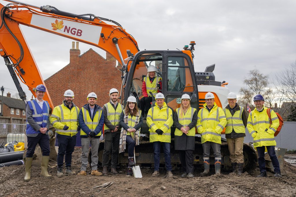 9 men and 1 woman are standing in a row on top of a mound of earth on a building site. Behind them is a digger with a woman sat in it. Everyone is wearing hi-viz and hard hats.