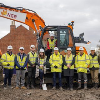 9 men and 1 woman are standing in a row on top of a mound of earth on a building site. Behind them is a digger with a woman sat in it. Everyone is wearing hi-viz and hard hats.
