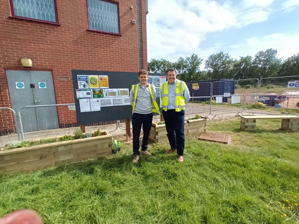 Two men in hi-viz jackets stand on a grassy garden area with planters and benches in front of a red-brick building.