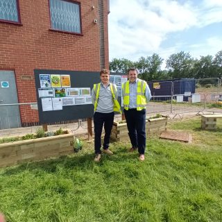 Two men in hi-viz jackets stand on a grassy garden area with planters and benches in front of a red-brick building.