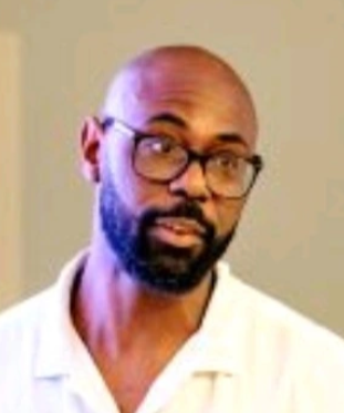A headshot of a man with a short dark beard, brown eyes and is bald. He is wearing glasses and a white open-collar shirt.