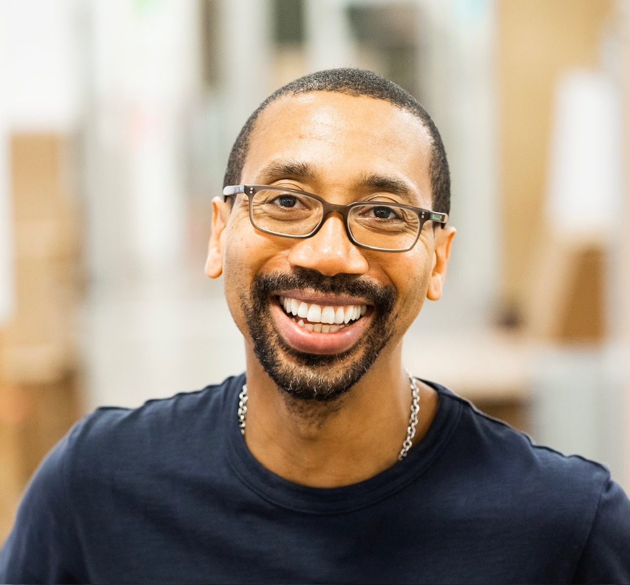 A headshot of a man with a short dark beard, brown eyes and is bald. He is wearing glasses and a white open-collar shirt.
