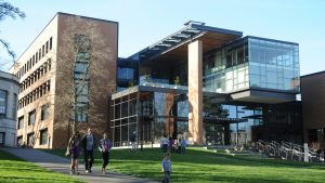 A picture of the outside of a university building. It's a brown brick and glass building with a sloping lawn at the front. Students are walking down a pathway and sat on the lawn.