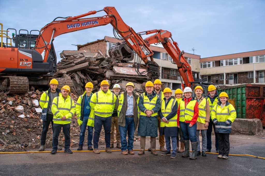 A group of 11 men and 3 women in hard hats and hi-vis vests are grouped together on a building site. They are standing in front of two plant vehicles which are on top of rubble. 1970s style block of flats are in the background.