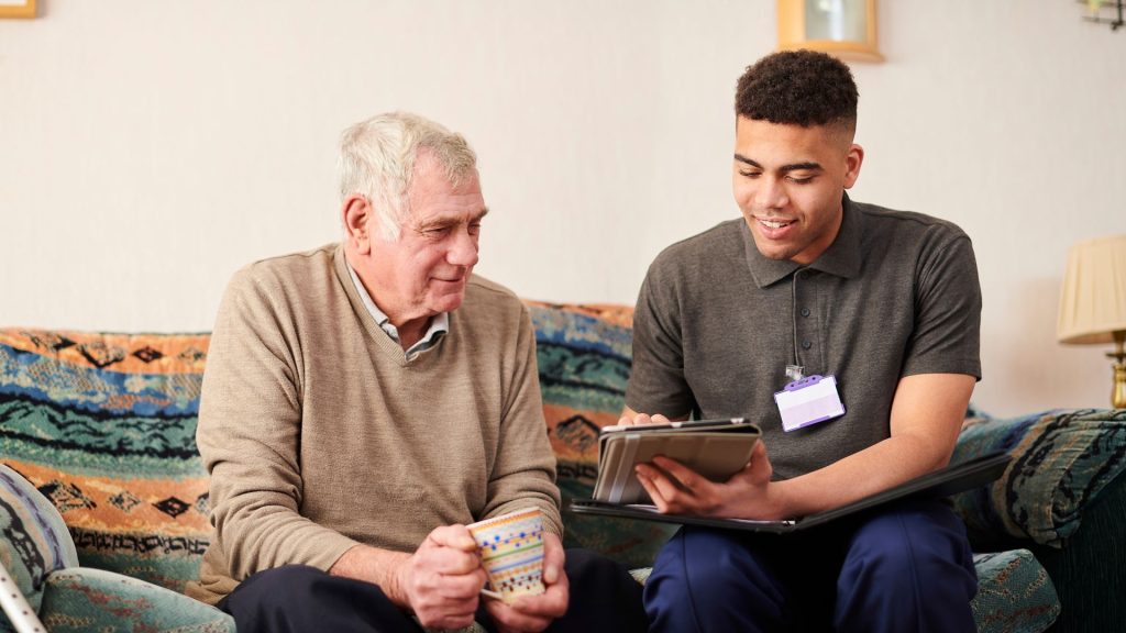Two men are sat on a sofa. One is elderly with short white hair and is wearing a beige jumper with a grey collared shirt. He is holding a mug in both hands. He is looking at the ipad that the other man is holding. The other man is younger and has short black curly hair. He is wearing a grey short-sleeved polo shirt with an ID badge clipped to it. He has a black folder open on his lap and is showing the older man something on an ipad.