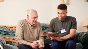 EDI-webinar-image Two men are sat on a sofa. One is elderly with short white hair and is wearing a beige jumper with a grey collared shirt. He is holding a mug in both hands. He is looking at the ipad that the other man is holding. The other man is younger and has short black curly hair. He is wearing a grey short-sleeved polo shirt with an ID badge clipped to it. He has a black folder open on his lap and is showing the older man something on an ipad.