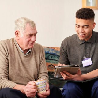 Two men are sat on a sofa. One is elderly with short white hair and is wearing a beige jumper with a grey collared shirt. He is holding a mug in both hands. He is looking at the ipad that the other man is holding. The other man is younger and has short black curly hair. He is wearing a grey short-sleeved polo shirt with an ID badge clipped to it. He has a black folder open on his lap and is showing the older man something on an ipad.