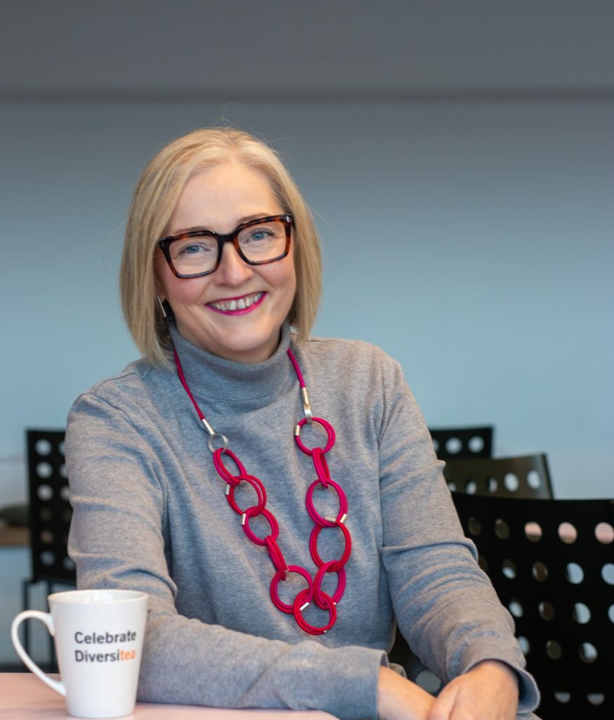 A headshot of a woman with a long blonde bob and thick framed glasses is smiling at the camera. She is wearing a grey turtle neck jumper and a large chunky red chain necklace.
