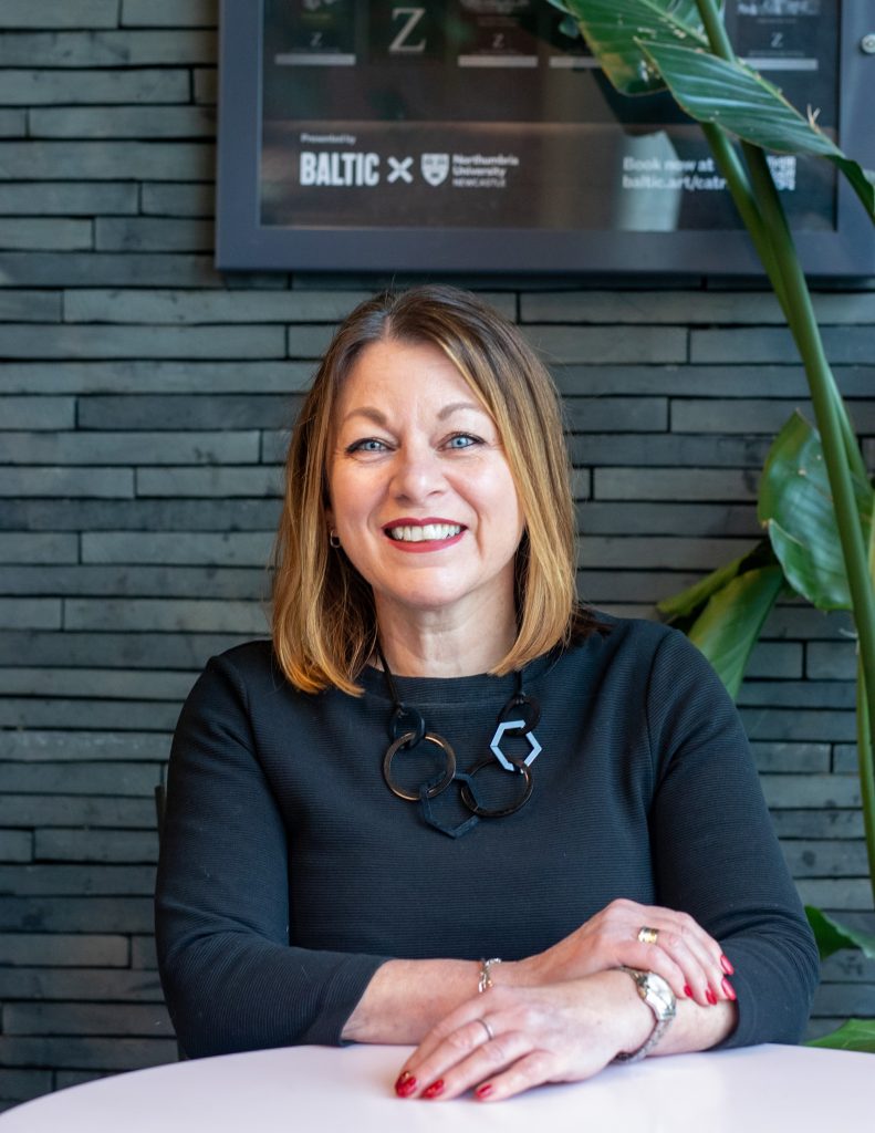 A  head and body shot of a woman with light brown hair and blue eyes. She is smiling at the camera. She is wearing a black jumper and chunky black necklace made up of ring and hexagon shapes. She is sat has her hands crossed in front of her. 