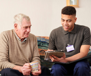 Two men are sat on a sofa. One is elderly with short white hair and is wearing a beige jumper with a grey collared shirt. He is holding a mug in both hands. He is looking at the ipad that the other man is holding. The other man is younger and has short black curly hair. He is wearing a grey short-sleeved polo shirt with an ID badge clipped to it. He has a black folder open on his lap and is showing the older man something on an ipad.