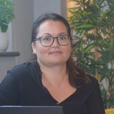 A woman with brown eyes and dark hair that is tied in a ponytail is sat at her laptop. She has glasses and is wearing a black blouse.