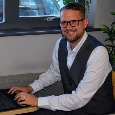 A man with short brown hair, short beard and glasses. He is wearing a white shirt, grey waistcoat and is typing on a laptop. His face is turned to the camera and is smiling.