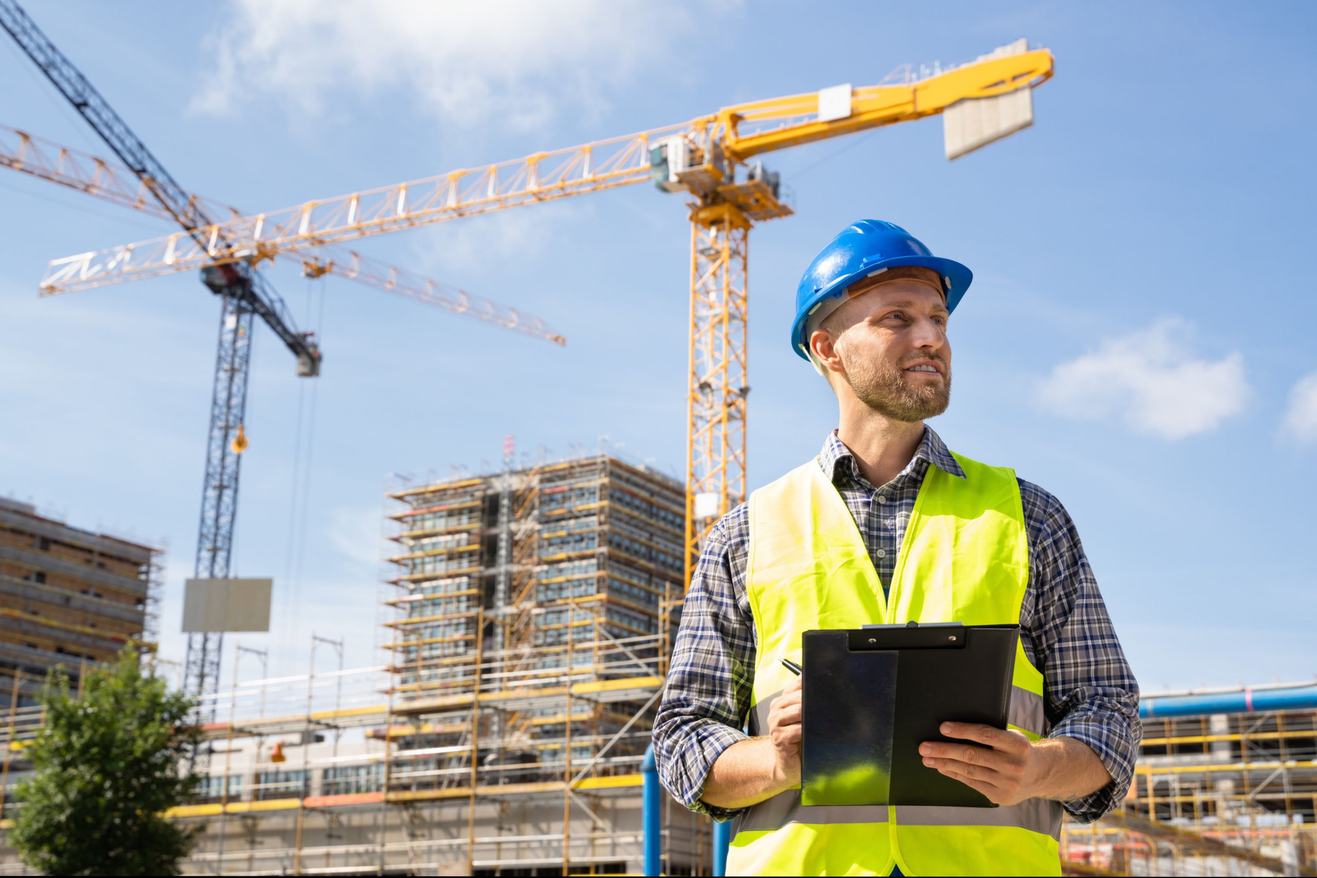 A Construction worker with a hard hat, high-viz and clipboard is stood in front of a building covered in scaffolding. There are cranes in the background.
